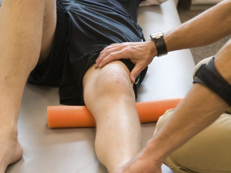 Bild einer Kniebehandlung in der Physiotherapie. Der Therapeut umgreift mit seinen Fingern die Kniescheibe des Patienten